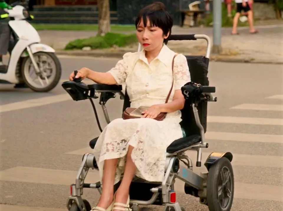 Cảnh phim ĐỢI: người phụ nữ mặc váy ren trắng ngồi xe lăn điện ở vạch sang đường, nhìn về phía trước
