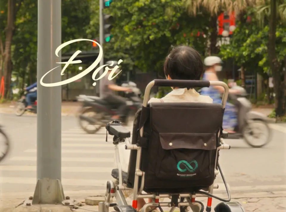 Cảnh phim ĐỢI: người phụ nữ ngồi xe lăn điện chờ ở vạch sang đường, chữ Đợi hiện trên khung hình